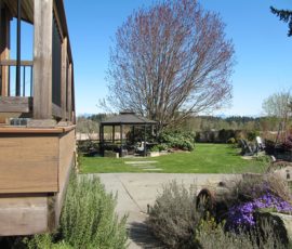 Deck with wooden railing overlooks a grassy yard with a gazebo, large tree, and various plants under a clear blue sky.