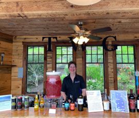 A person stands behind a wooden bar with drinks, cans, bottles, and signs displayed on the counter.