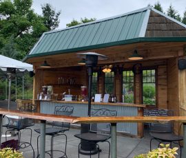 Outdoor wooden bar with stools, a green metal roof, and surrounding greenery on a cloudy day.