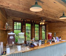 Outdoor wooden drink stand with large beverage dispensers, cups, snacks, and bottled drinks on the counter.