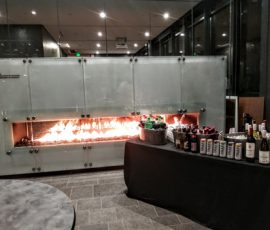 A modern indoor fireplace with a table set for drinks and bottles in a sleek, glass-walled room.