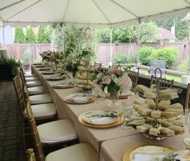 Long table set for an outdoor event under a tent, decorated with flowers, plates, and a tiered tray of finger sandwiches.