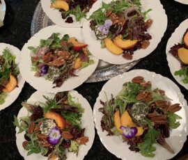 Plates of mixed green salad with pecans, peach slices, cheese, and edible flowers on white dishes.