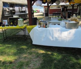 Outdoor refreshment table with drinks, glassware, and a dispenser set up next to a food truck and shaded pavilion.