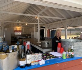 A woman stands behind a counter with bottles, cans, and drinks in an open, white-painted outdoor pavilion.