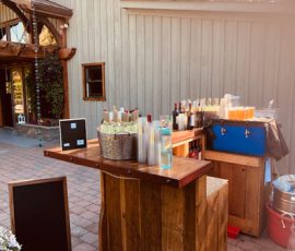 Outdoor wooden bar setup with assorted bottled drinks, plastic cups, a cooler, and chalkboard signs on a patio.