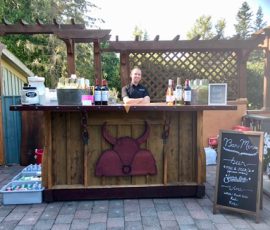 Outdoor bar with a bartender standing behind it, wine bottles on display, and a chalkboard menu on the side.